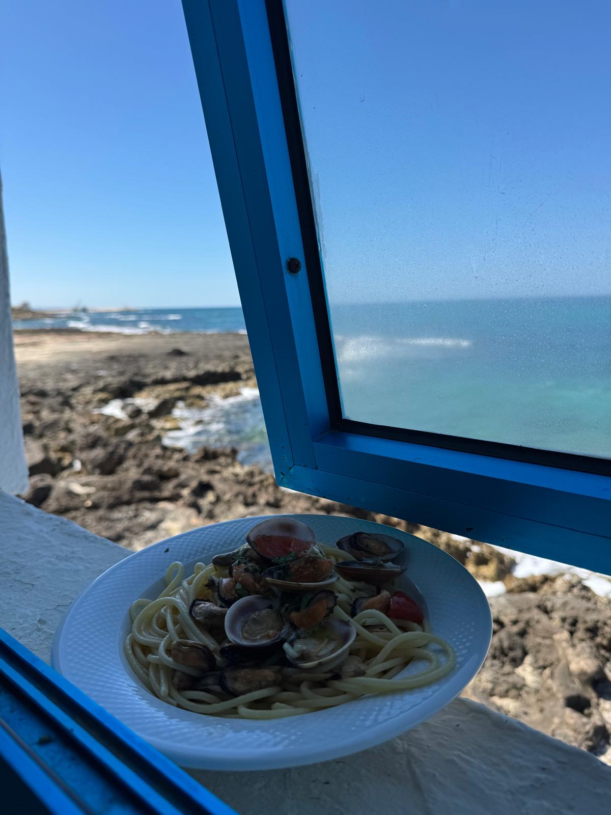 A plate of pasta with clams beside an open blue window overlooking the sea
